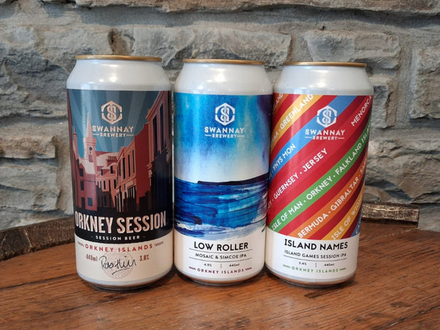 Three beer cans with different designs on a wooden surface against a stone wall background